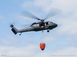 Zdjęcie przedstawia znajdujący się w locie helikopter Black Hawk z przytwierdzonym pomarańczowym koszykiem typu Bambi Bucket.