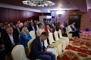 Zdjęcie przedstawia uczestników konferencji siedzących na krzesłach z białymi narzutami, które zostały ustawione w pięciu rzędach.