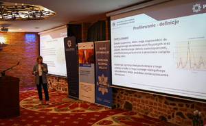 Zdjęcie przedstawia jedną z prowadzących konferencję. Za jej plecami znajduje się zestaw roll-upów promocyjnych, ona sama stoi za podium i zwraca się do swojej lewej strony, kierując wzrok na wyświetlacz, na którym ukazuje się prezentacja dotycząca &amp;quot;Profilowania (wyświetlony jest napis: Profilowanie - definicja oraz treść prezentacji).