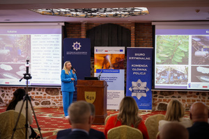 Zdjęcie przedstawia jedną z prowadzących konferencję, która ubrana jest jasnoniebieski kombinezon. Za jej plecami znajduje się zestaw roll-upów promocyjnych. Po jej lewej i prawej znajdują się wyświetlacze, na których prezentowana jest ta sama prezentacja dotycząca kokainy.
