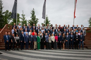 Zdjęcie przedstawia zbiorową fotografię uczestników spotkania. Zostało wykonane na zewnątrz hotelu. Niebo jest zachmurzone, z tyłu uczestników znajdują się drewniane schody, prowadzące do tarasu, na którym ustawione zostały ławki.
W tle widać pojedyncze drzewa iglaste oraz maszty z nie do końca rozwiniętymi flagami.
