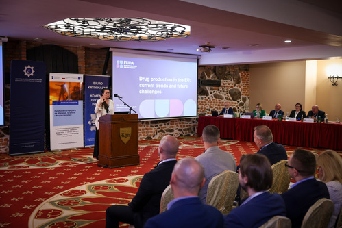 Zdjęcie przedstawia jedną z prowadzących konferencję. Za jej plecami znajdują się trzy roll-upy promocyjne, obok których widać wyświetlacz, na którym pokazuje się napis na niebieskim tle: &quot;Drug production in the EU: current trends and future challenges&quot;.
