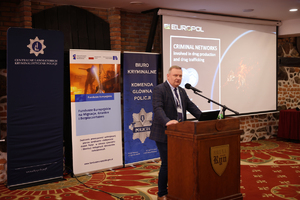 Zdjęcie przedstawia jednego z prowadzących konferencję, który stoi za podium i mówi do mikrofonu, za jego plecami znajdują się roll-upy promocyjne, obok których stoi wyświetlacz, na którym ukazana jest prezentacja dotycząca &quot;Criminal Networks&quot;.