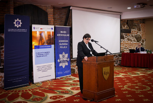 Zdjęcie przedstawia jedną z prowadzących konferencję, stoi ona za podium, patrzy na kartkę. Za nią ustawione zostały trzy roll-upy promocyjne oraz wyświetlacz.