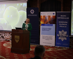 Zdjęcie przedstawia jedną z prowadzących konferencję. Za jej plecami ustawione zostały trzy roll-upy promocyjne, obok których stoi wyświetlacz, na którym ukazana została prezentacja.
