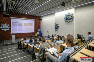 Zdjęcie przedstawia rzut na salę z perspektywy uczestników spotkania. Centralne miejsce zajmuje tablica, na której wyświetlono informacje o spotkaniu.