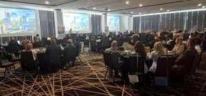 Zdjęcie przedstawia siedzących na sali uczestników konferencji, którzy obserwują wyświetlaną na dwóch ekranach prezentację.