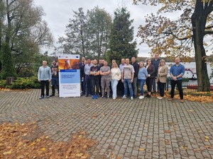 Zdjęcie przedstawia uczestników szkolenia realizowanego w ramach projektu DISLABOR. Uczestnicy pozują na przestrzeni otwartej, za plecami mają jeziora i drzewa. Obok grupy znajduje się roll-up projektowy.