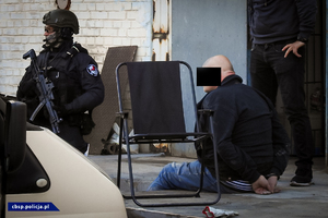 Policyjny antyterrorysta z zatrzymanym siedzącym na ziemi. Obok stojący policjant.