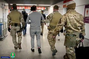Funkcjonariusze prowadzą mężczyznę w kajdankach założonych na ręce i nogi po terenie lotniska.