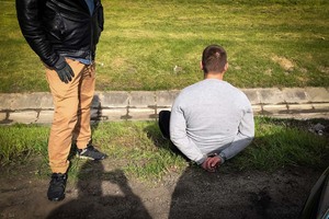 Mężczyzna w kajdankach siedzący na trawniku. Obok stoi policjant w stroju cywilnym.