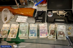 Liczne banknoty o nominałach od 10 do 500 PLN ułożone w stosach. Broń palna leżąca w walizeczce.