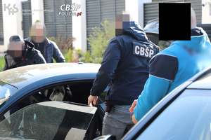 Funkcjonariusze CBŚP dokonują przeszukania samochodu. Obok stojący zatrzymany w kajdankach na rękach.