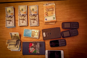 Banknoty o nominałach 100 USD, 50 EUR oraz 100 PLN, a także telefony komórkowe i wagi elektroniczne leżące na blacie stołu.