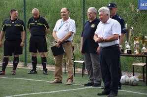 Fotografia kolorowa przedstawiająca boisko. Po lewej stronie widać dwóch sędziów piłkarskich. W dalszej kolejności przedstawicieli IPA oraz Burmistrza Pruchnika, który wita zgromadzone drużyny.