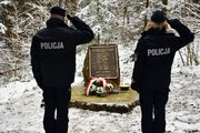 Złożenie kwiatów i zapalenie zniczy przy obelisku upamiętniającym katastrofę śmigłowca w Cisnej przez policjantów. Policjant i policjantka (widok zza ich pleców) oddają honory przed obeliskiem.