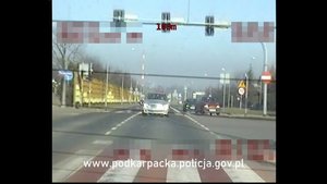 Zdjęcie z policyjnego wideorejestratora - widok z tylnego okna. Na pierwszym planie srebrny samochód jadący za policyjnym radiowozem, przejeżdżający przez skrzyżowanie na wprost. W tle inne pojazdy. Na dole ekranu napis www.podkarpacka.policja.gov.pl.