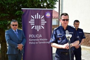 I Zastępca Komendanta Wojewódzkiego Policji w Rzeszowie stojący przed mikrofonem, za nim stoi komendant miejski z Tarnobrzega i Wójt Gminy Grębów