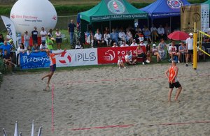 na piaszczystym boisku dwóch zawodników rozgrywa mecz siatkówki plażowej, poza boiskiem, pod namiotami siedzą widzowie