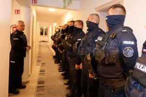 Po lewej Komendant Wojewódzki Policji w Rzeszowie w czasie odprawy na korytarzu. Po prawej w szeregu stoją policjanci, na twarzach mają kominy lub kominiarki.