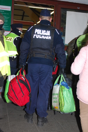 Zdjęcie kolorowe przedstawia policjanta który pomaga uchodźcom z Ukrainy  w przenoszeniu bagaży