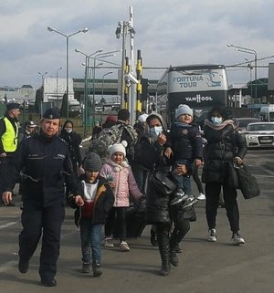 Zdjęcie kolorowe przedstawia policjanta który pomaga uchodźcom z Ukrainy  w przenoszeniu bagaży i przenoszą dzieci