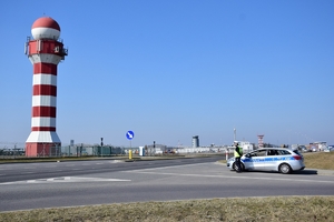 Policjant ruchu drogowego stoi przy radiowozie (po prawej stronie) przy wyjeździe z bocznej ulicy. W tle budynki i infrastruktura lotniska.