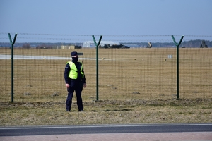 Policjant oddziału prewencji stoi przy ogrodzeniu lotniska. W tle pas startowy.