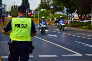 Na pierwszym planie (po lewej) policjant stojący na ulicy, w tle na środku dwaj policyjni motocykliści jadący na sygnałach.