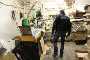 Policjant urany w czarną rękawiczkę z białym napisem policja stoi tyłem przy maszynie wykorzystywanej do produkcji podrabianych papierosów
