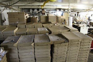 Na zdjęciu widać rzędy kartonów z papierosami, w tle pomieszczenia fabryki z maszynami wykorzystywanymi do produkcji podrabianych papierosów
