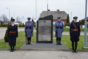 Policjanci w mundurach wyjściowych z bronią przy nodze, stoją przy pamiątkowym obelisku. Po lewej i prawej stronie stoją policjant i policjantka w mundurze z czasów II RP.
