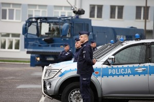 Stojący przy oznakowanych radiowozach umundurowaniu policjanci, a w tle niebieski duży samochód - policyjna polewaczka.