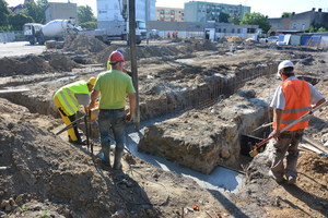 Robotnicy stoją tyłem i patrzą na prace budowalne - dziury w ziemi.