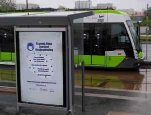Plakaty Krajowej Mapy Zagrożeń Bezpieczeństwa na przystanku komunikacji miejskiej. w Tle stoi tramwaj.