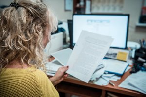 Kobieta siedząca i czytająca dokument, w tle monitor komputera i dokumentacja