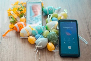 Dwa telefony komórkowe, na wyświetlaczu pierwszego połączenie do Babci Zosi, na drugim zdjęcie starszej kobiety. Tłem są wielkanocne, kolorowe jajeczka