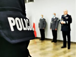 Na pierwszym planie napis policja na mundurze, w tle Komendant Wojewódzki Policji w Katowicach podczas przemówienia