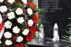 Na pierwszym planie wieniec z biało-czerwonymi kwiatami, na dalszym planie zapalone znicze oraz fragment pomnika z kilkoma nazwiskami tragicznie zmarłych osób.