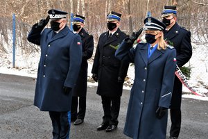 Komendant Wojewódzki Policji wraz z Panią Naczelnik Gabinetu Komendanta Wojewódzkiego Policji oddają honor, w tle kilkuosobowa delegacja ze straży pożarnej.