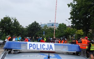 Kolorowa fotografia przedstawia radiowóz policyjny, w oddali widoczni rowerzysci.