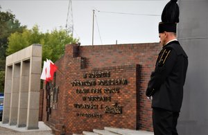 Kolorowe zdjęcie przedstawia tablice upamiętniająca śmierć górników. Na pierwszym planie górnik w mundurze, w tle widoczne flagi Polski.