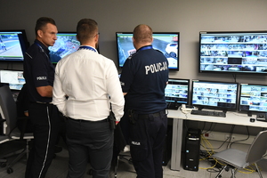 Stanowisko dowodzenia w Arenie Gliwice. Dwaj umundurowani policjanci oraz mężczyzna w białej koszuli obserwują monitoring.