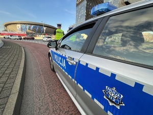 Zdjęcie kolorowe: na pierwszym planie oznakowany radiowóz, przed nim widoczni policjanci. W tle widoczny budynek katowickiego spodka.