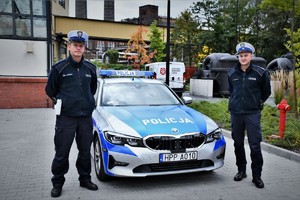 Na zdjęciu dwaj umundurowani policjanci i radiowóz na placu