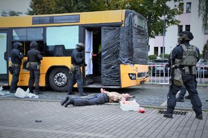 Zdjęcie. Widoczni umundurowani policjanci z bronią maszynową przed autobusem, oraz osoba leżąca na chodniku