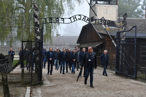osoby przechodzące przez bramę obozu Auschwitz z napisem Arbeit Macht Frei.  W tle budynki obozu.