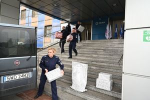 Policjanci znoszą spakowane paczki do samochodu przed komendą