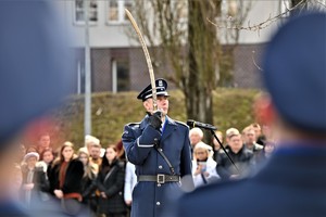 zdjęcie przedstawia dwóch policjantów stojących tyłem, między nimi widoczny salutujący szablą dowódca uroczystości