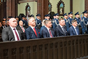 kolorowe zdjęcie przedstawiające mężczyzn siedzących w ławce bazyliki, w tle stoją policjanci kompani honorowej